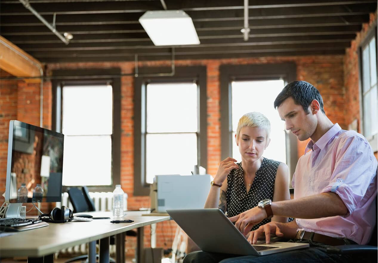 A portable ADT panic button device clipped onto the belt of a person wearing business-casual clothes. Two business owners in a modern office review information on a laptop, illustrating video business operations with ADT security.